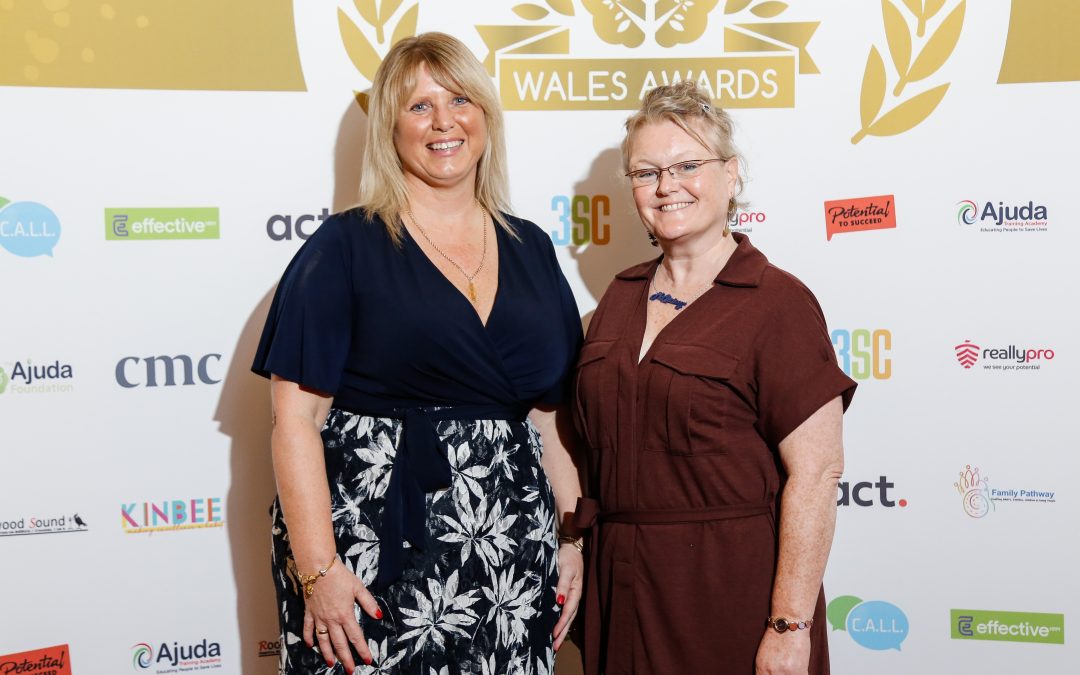 Two women standing in front of a presentation board at an awards ceremony. They look very smart, and are smiling directly at the camera lens. One woman is blonde, and wearing a black and white dress. This is Dawn Evans, CEO of the Ajuda Foundation. The other is a greying redhead. She's wearing a brown dress with a tie belt. This is Bobbie Allen, and she's the project co-ordinator for the Ajuda Foundation.