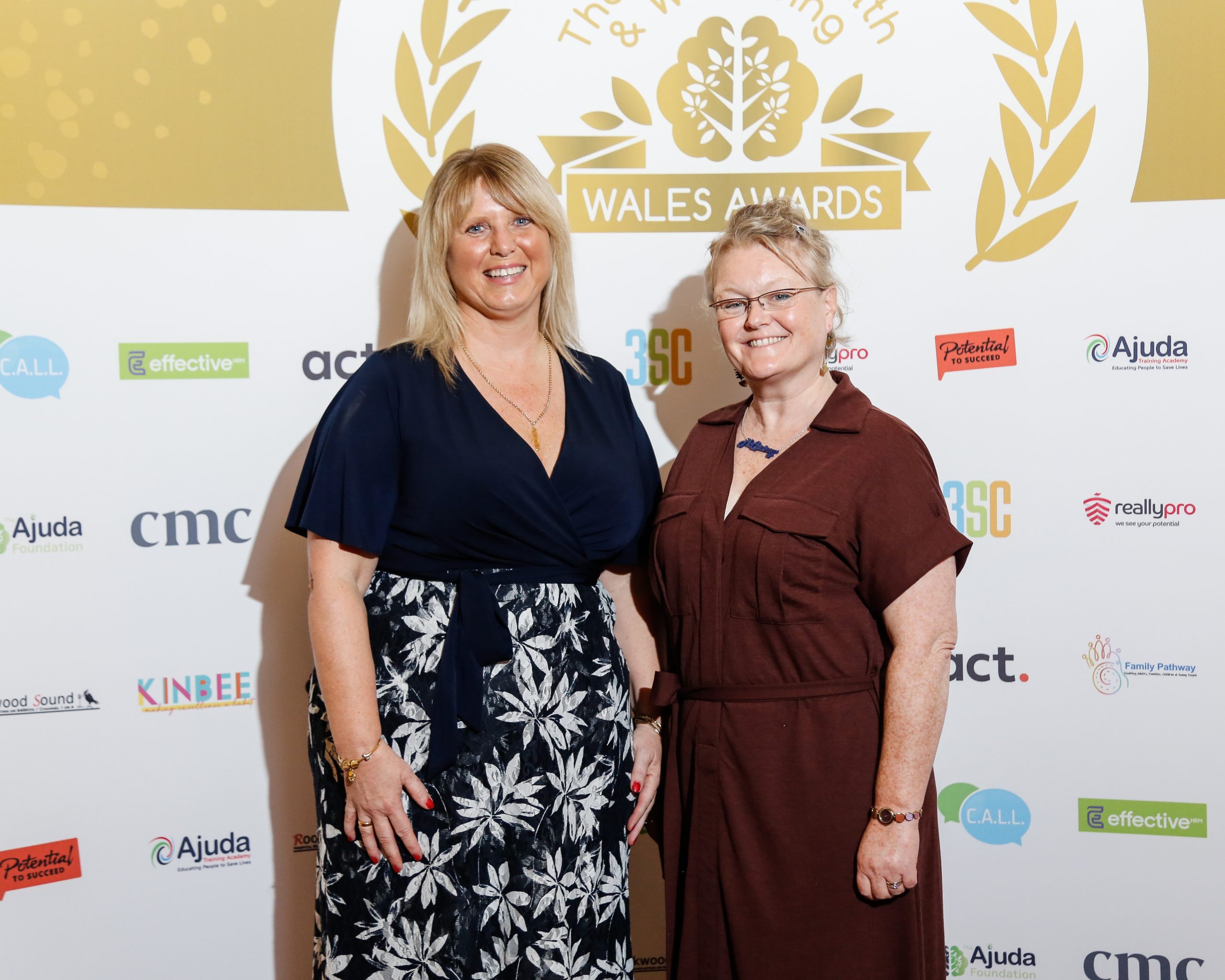 Two women standing in front of a presentation board at an awards ceremony. They look very smart, and are smiling directly at the camera lens. One woman is blonde, and wearing a black and white dress. This is Dawn Evans, CEO of the Ajuda Foundation. The other is a greying redhead. She's wearing a brown dress with a tie belt. This is Bobbie Allen, and she's the project co-ordinator for the Ajuda Foundation.